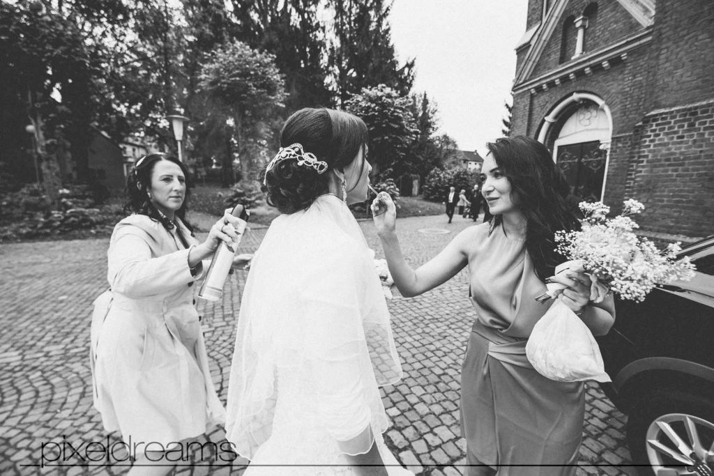 Hochzeit Fotograf Köln– Brautjungfern verpassen der braut noch ein letztes brautstyling bevor sie in die kirche gehen. haarspray und lipgloss sind ihre werkzeuge