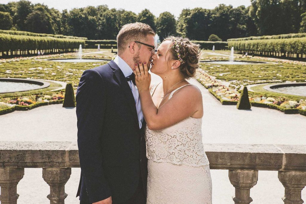 Brautpaar küsst sich auf Söller mit Schlossgarten im Hintergrund. Medium Shot/ Naheinstellung)