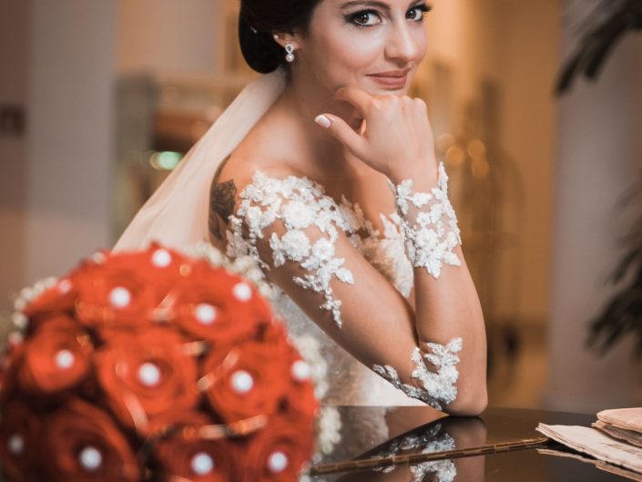 Hochzeitsfoto einer türkischen Braut Mit rotem Rosestrauß Bouquet im Vordergrund. Braut schaut lächelnd und verlegen in die Kamera. Perfekte Brautfrisur und tolles perfektes Braut Mac ab. Sie posiert über einem Klavier.