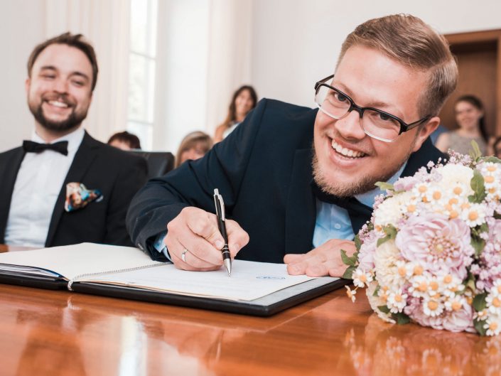 Hochzeitsfoto vom Bräutigam während der standesamtlichen Zeremonie. Bräutigam unterschreibt seine Heiratsurkunde Und Blick glücklich in die Kamera. Kreatives Hochzeitsbild eines professionellen Hochzeitsfotografen.