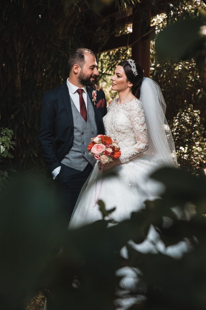 Hochzeitsfoto eines Brautpaares im japanischen Garten in Leverkusen. Hochzeitsfotograf steht hinter Ästen und beobachtet das Paar aus einer Voyeur-Perspektive. Kreatives Hochzeitsfoto.