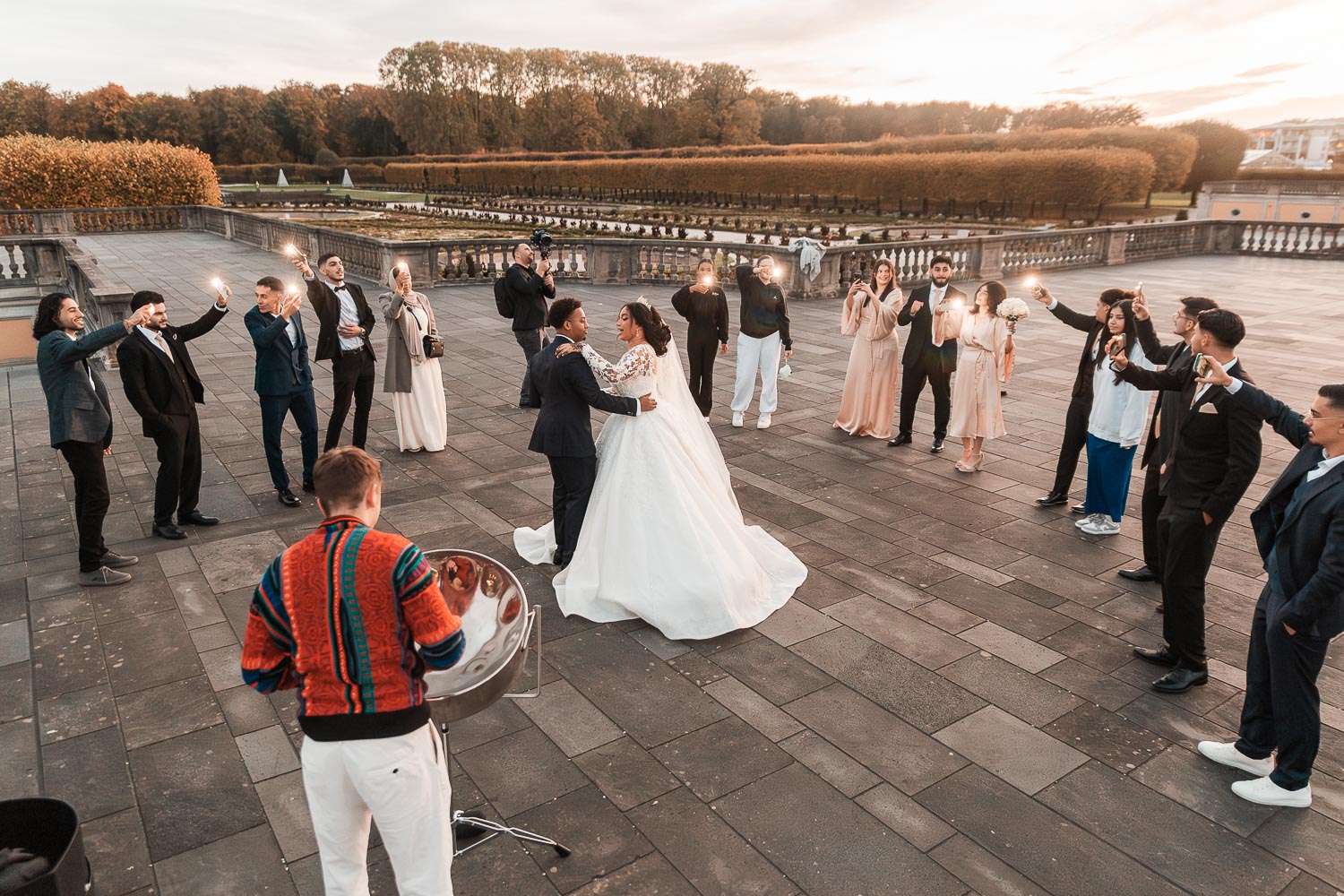 Ein Steeldrum Straßenmusiker spielt moderne Musik, während das Brautpaar von ihren Freunden umgeben tanzt. Die Szene spielt sich auf der Terrasse des Schlosses in Augustusburg ab, im Hintergrund ist der Schlossgarten zu sehen.