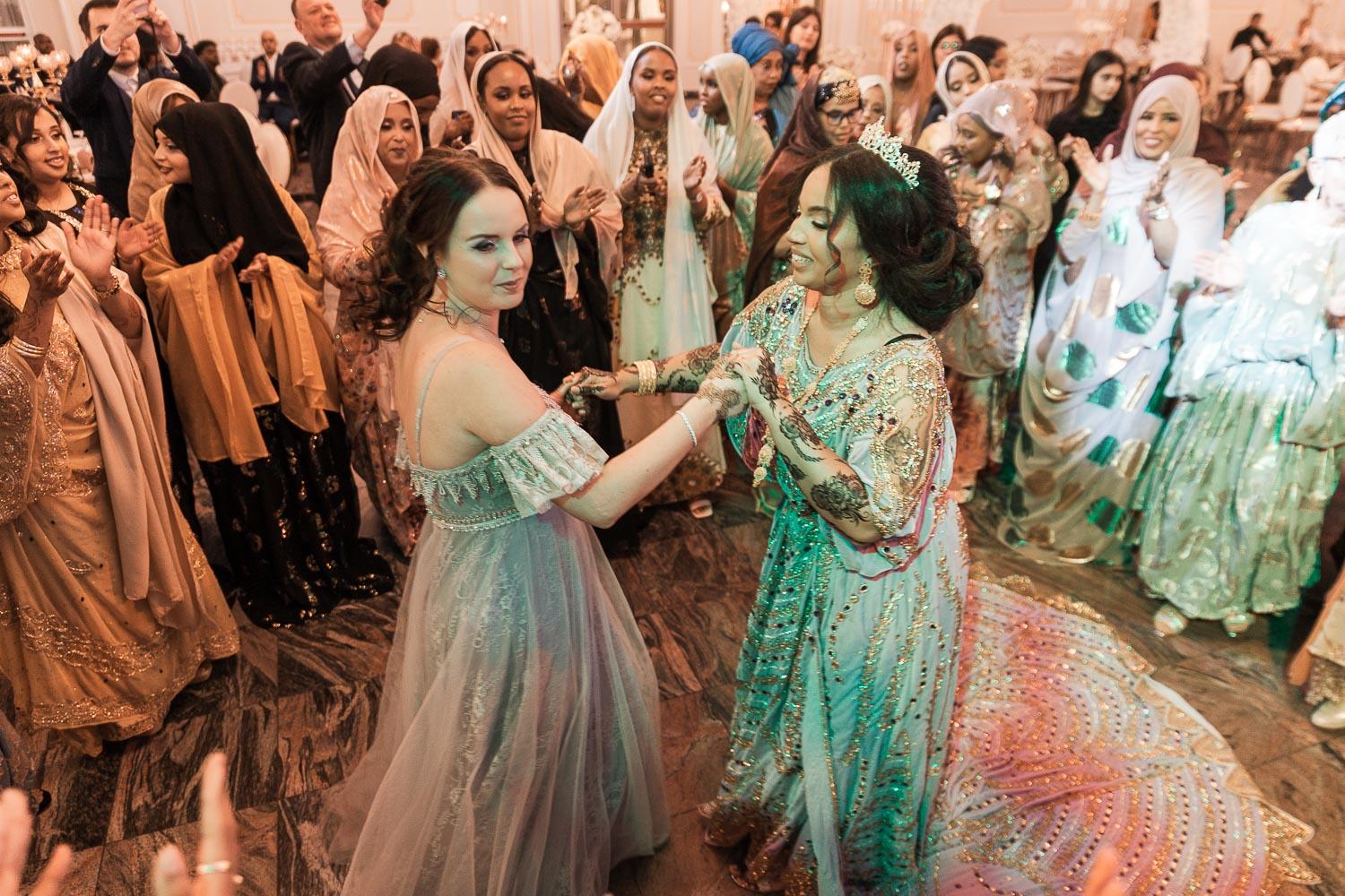Eine somalische Braut in traditioneller Kleidung tanzt mit ihren Brautjungfern, während im Hintergrund andere Gäste sich bei einer Hochzeit im Event Palast Hürth amüsieren. Frauen tragen traditionelle somalische Kleidung Diraa/Guntiino/Garbasaar.