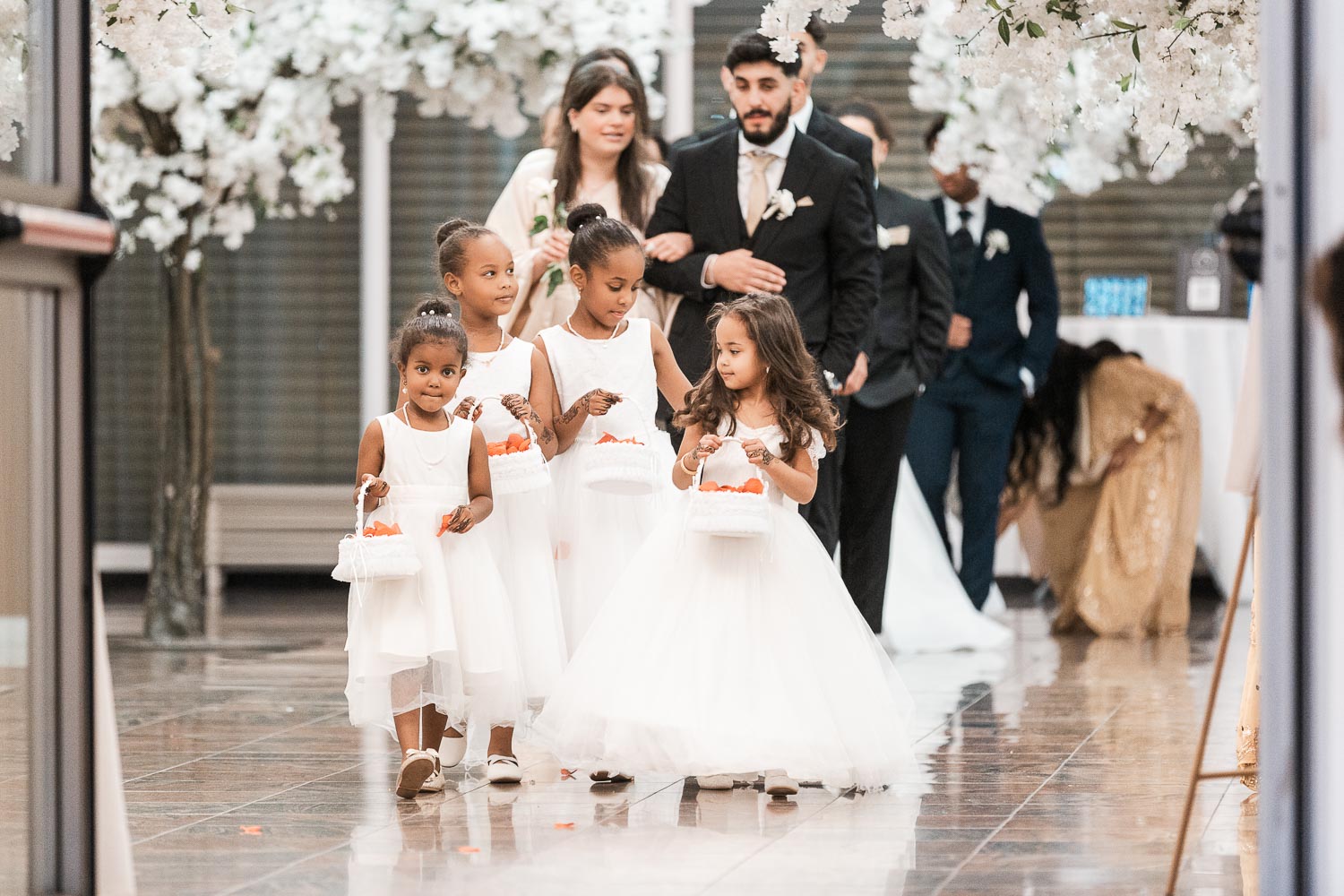 Vier hübsche Blumenmädchen in weißen Kleidern schreiten mit Blumenkörbchen voran und betreten den Event Palast Hochzeitssaal und werden begleitet von Trauzeugen.
