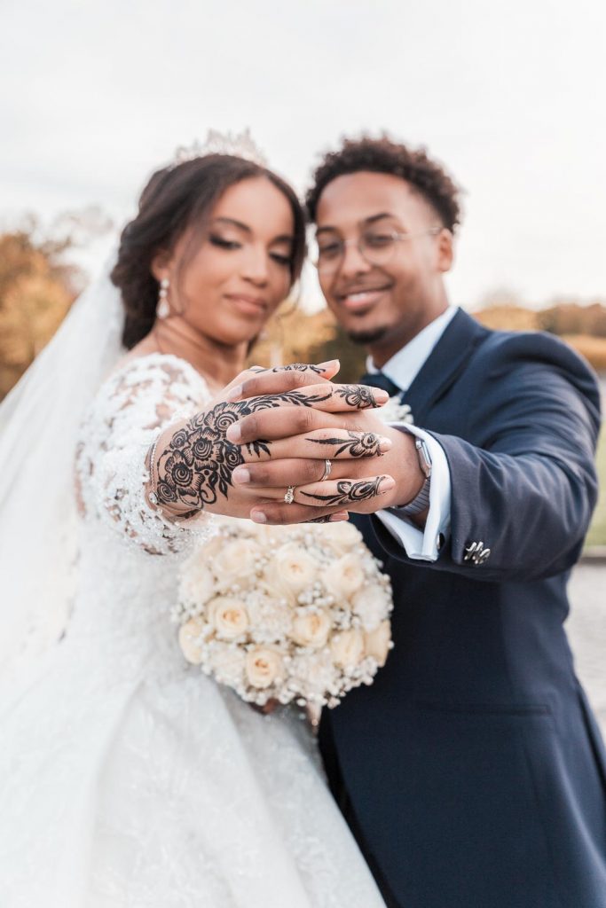 Hochzeitsfotograf Bruehl Schloesser - somalisch afrikanisch hochzeit - Hochzeitsreportage_2_Brautpaar Shooting Hände des Brautpaares sind ineinander verschränkt und zeigen Hennatatoo und Eheringe Brautstrauß unscharf im Hintergrund sichtbar