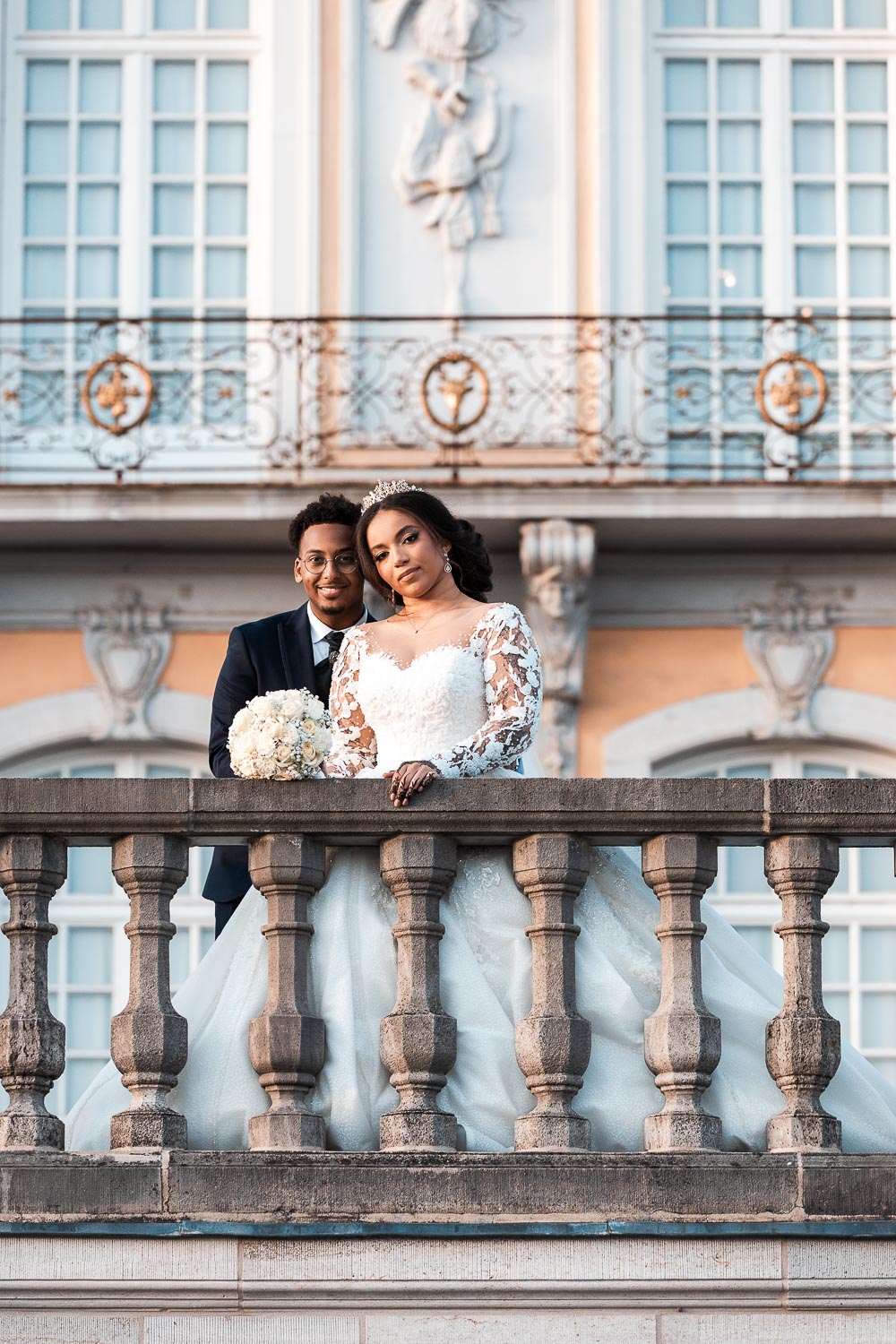 Brautpaar posiert für das Hochzeitsshooting am Schloss Augustusburg in Brühl auf der Terrasse. Die halb afrikanische Braut hält den Brautstrauß mit weißen Rosen in der Hand und legt es auf die Brüstung ab.