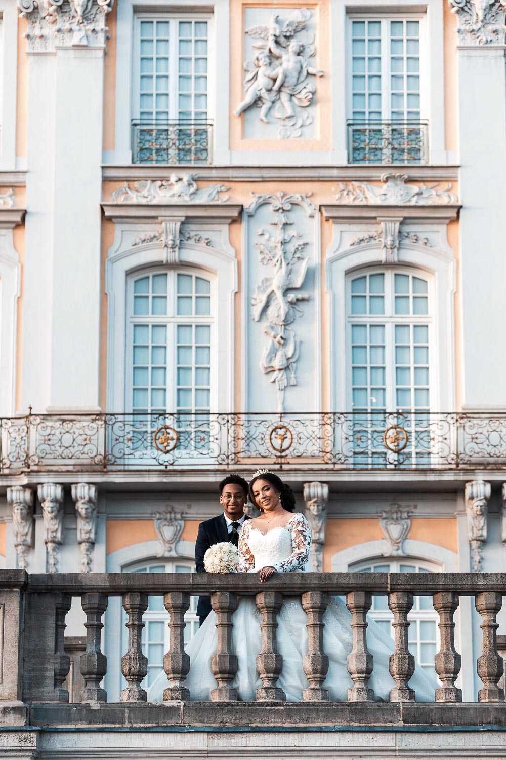 Brautpaar posiert für das Hochzeitsshooting am Schloss Augustusburg in Brühl auf der Terrasse. Die halb afrikanische Braut hält den Brautstrauß mit weißen Rosen in der Hand und legt es auf die Brüstung ab. In dieser Perspektive ist die Barockfassade des Schlosses leicht verschwommen im Hintergrund gut zu sehen.