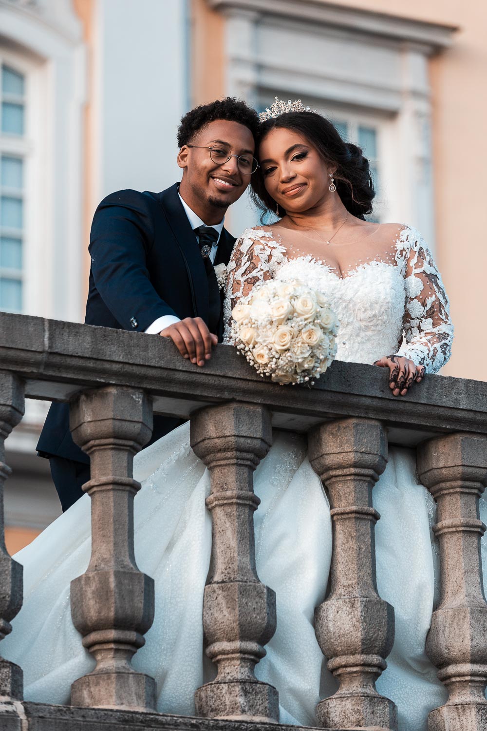 Brautpaar posiert für das Hochzeitsshooting am Schloss Augustusburg in Brühl auf der Terrasse. Die halb afrikanische Braut hält den Brautstrauß mit weißen Rosen in der Hand und legt es auf die Brüstung ab.