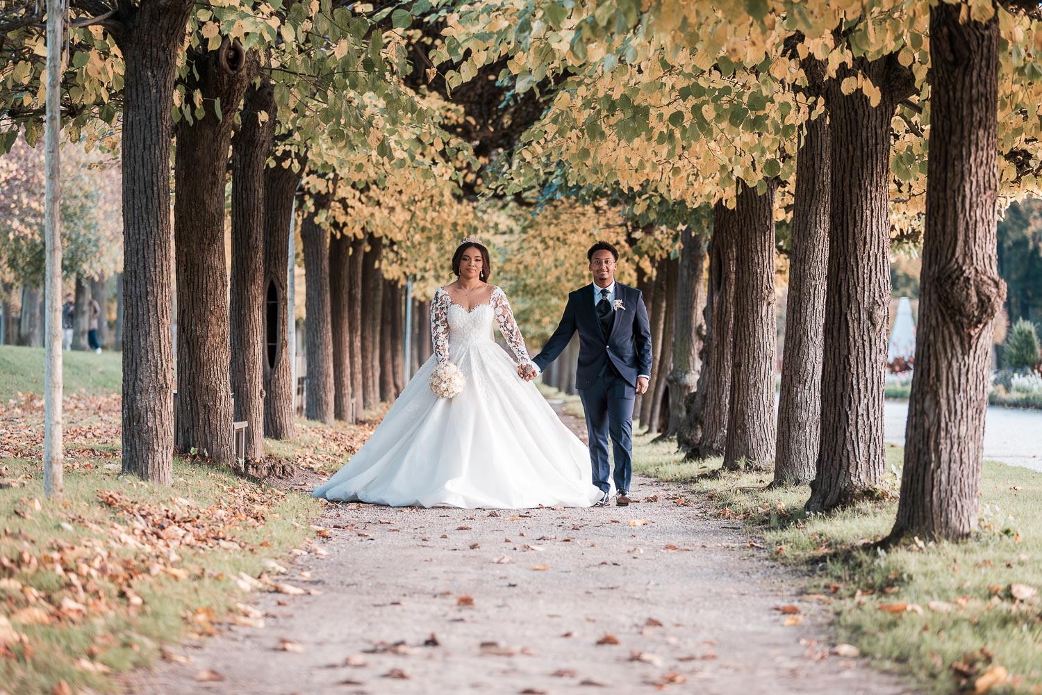Das hübsche Brautpaar, Hand in Hand, spaziert durch eine kleine Allee im Schlossgarten Augustusburg, die direkt auf den Hochzeitsfotografen zuführt. Von der Perspektive des Fotos aus betrachtet, befindet sich links parallel dazu eine größere Allee.