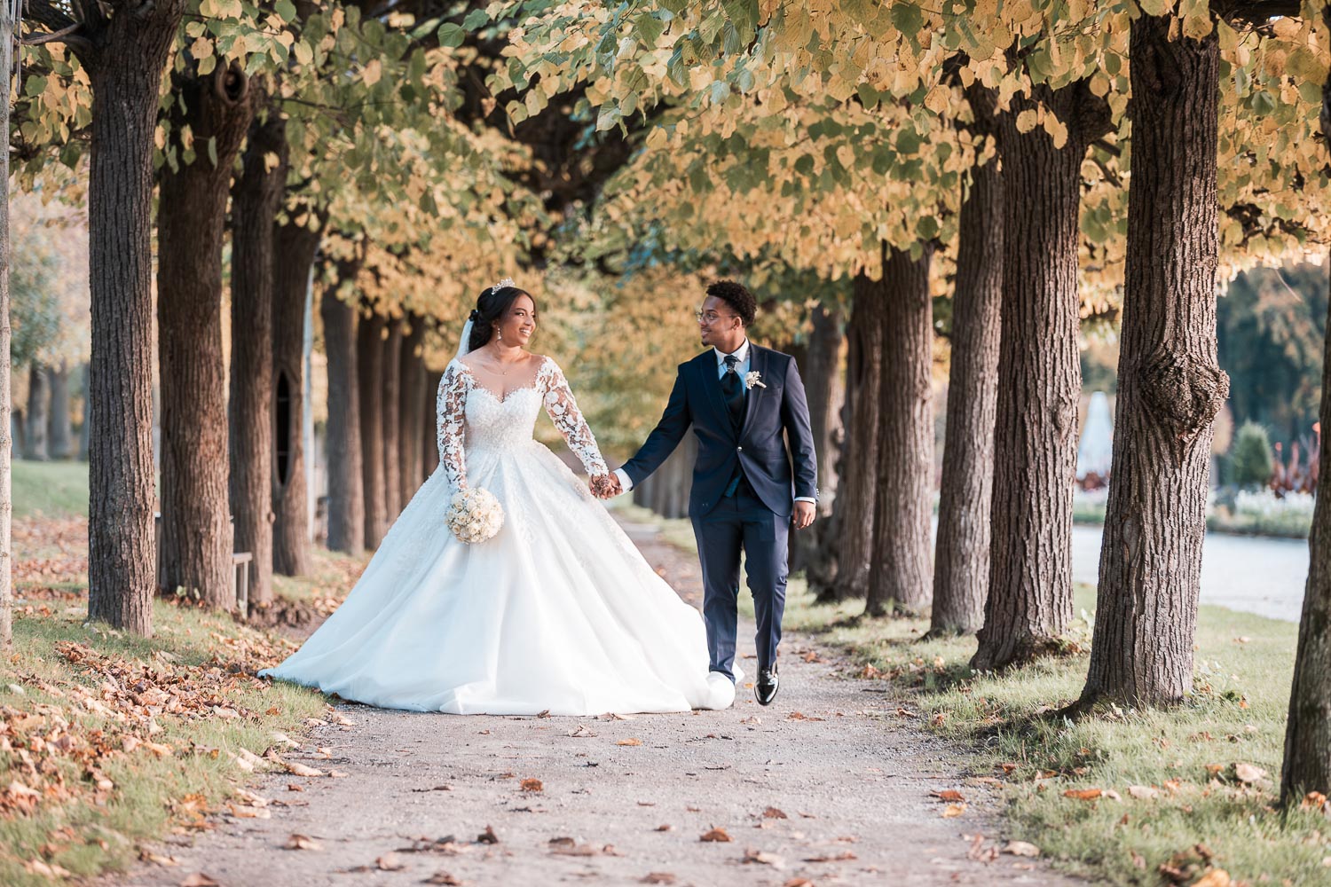 Das hübsche Brautpaar, Hand in Hand, spaziert durch eine kleine Allee im Schlossgarten Augustusburg, die direkt auf den Hochzeitsfotografen zuführt. Von der Perspektive des Fotos aus betrachtet, befindet sich links parallel dazu eine größere Allee.