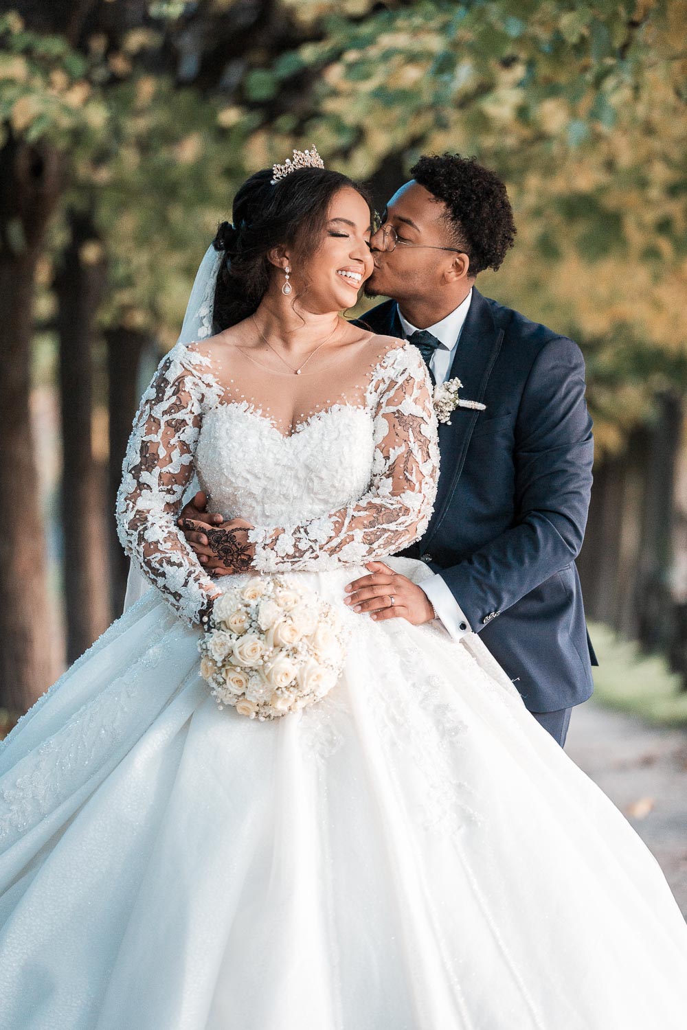 Der Bräutigam umarmt die Braut von hinten in der Allee und küsst sie auf die Wange, während sie lächelt. Das Hochzeitsfoto ist im amerikanischen Stil angeschnitten.