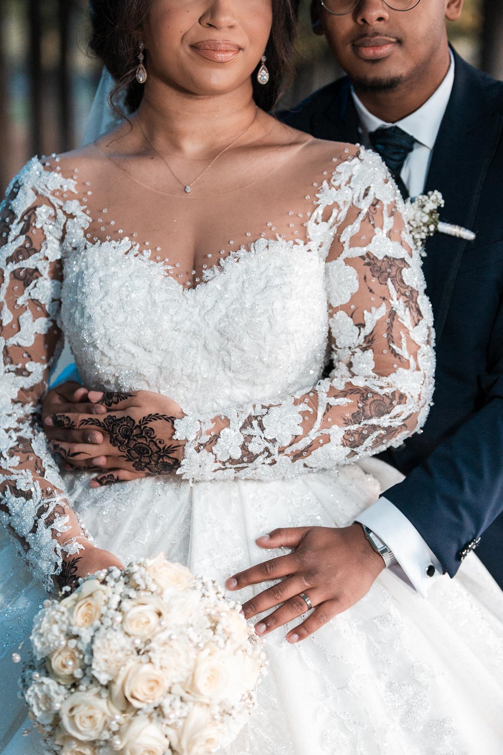 Der Bräutigam umarmt die Braut von hinten. Das Hochzeitsfoto ist unterhalb der Augen bis zur Hüfte angeschnitten und zeigt nur die Details des Kleides, von Hannah Tattoo, vom Braut Strauß, und Brautkleid und Anzug.