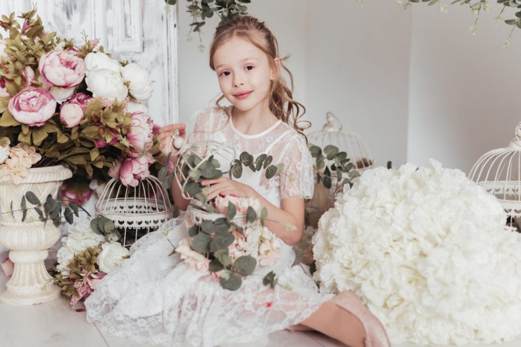 Ein bezauberndes Blumenmädchen sitzt zwischen vielen Blumengestecken und posiert für die Kamera des Hochzeitsfotografen