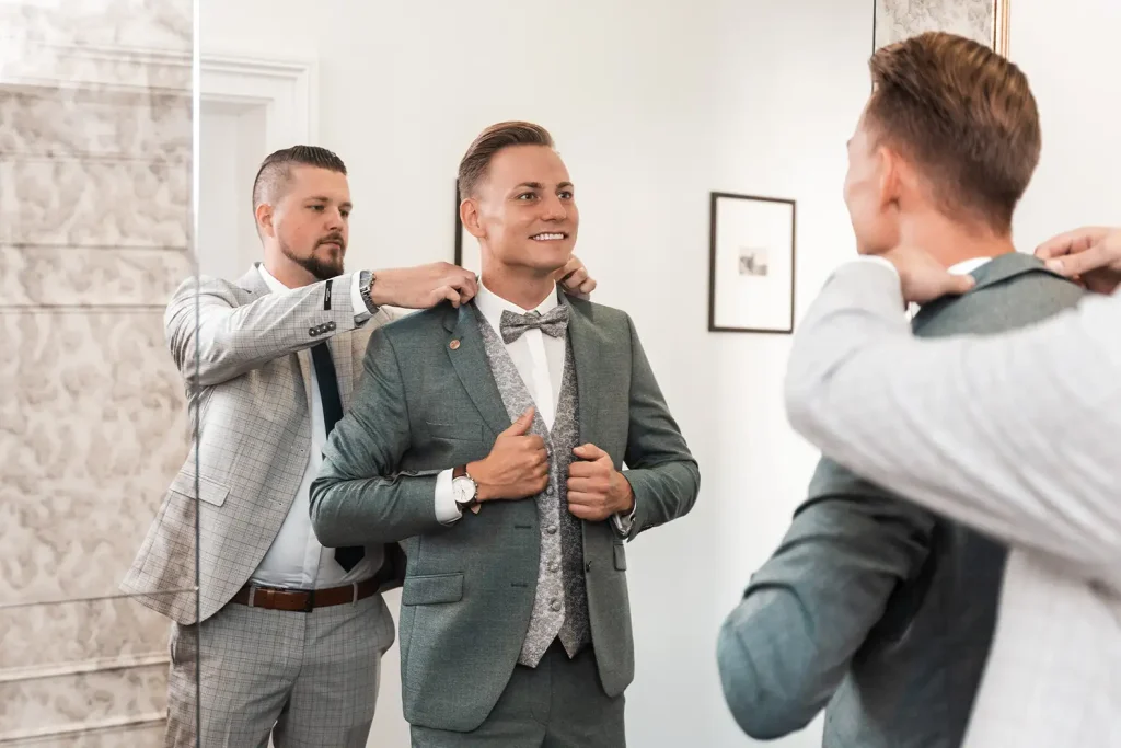 Bräutigam beim Getting Ready vor der Hochzeit, während sein Begleiter den Hochzeitsanzug richtet