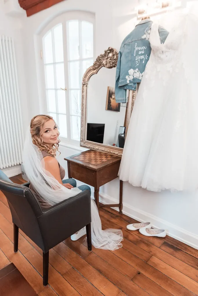 Braut mit Schleier beim Getting Ready neben ihrem Brautkleid vor der Hochzeit